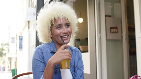 Young-biracial-woman-with-curly-blonde-hair-enjoys-a-drink-at-an-outdoor-cafe
