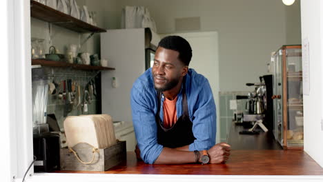 Un-Joven-Barista-Afroamericano-Se-Apoya-En-El-Mostrador-De-Una-Cafetería,-Sonriendo-A-La-Cámara