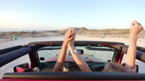 Two-people-raise-their-hands-joyfully-in-a-red-convertible-near-the-beach-on-a-road-trip