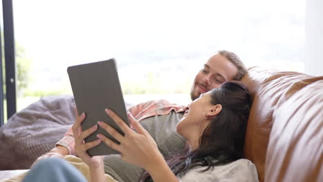 A-diverse-couple-relaxes-on-a-sofa-at-home,-using-a-tablet