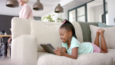 African-American-girl-lies-on-a-sofa-using-a-tablet,-her-smile-suggesting-enjoyment