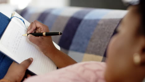 African-American-woman-writes-in-a-notebook,-seated-on-a-couch-at-home