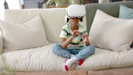 African-American-boy-wearing-a-virtual-reality-headset-sits-cross-legged-on-a-sofa-at-home
