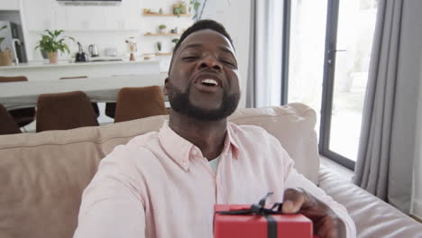 Afroamerikanischer-Mann-Mit-Roter-Geschenkbox-Lächelt,-Wirft-Einen-Kuss-In-Einem-Hellen-Wohnzimmer