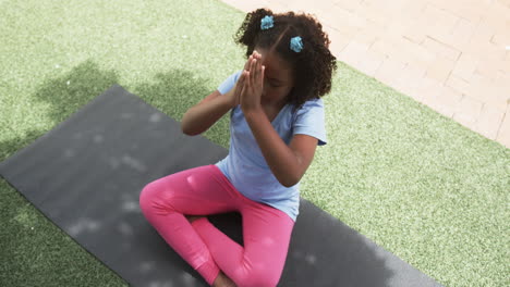 Biracial-girl-meditates-on-a-yoga-mat-outdoors-in-school,-hands-pressed-together