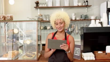 Young-biracial-female-barista-with-curly-blonde-hair-holds-a-tablet-in-a-bakery