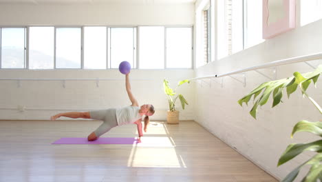 Caucasian-woman-holding-purple-ball,-doing-yoga-in-sunny-studio