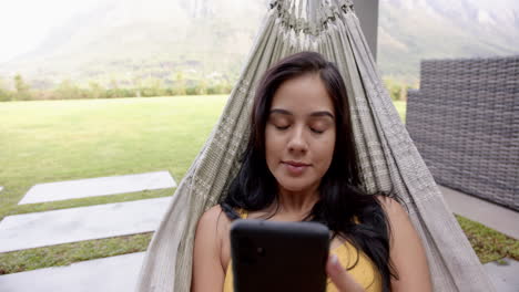 A-young-biracial-woman-relaxes-in-a-hammock-in-the-backyard-at-home,-smartphone-in-hand