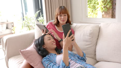 Zwei-Junge-Freundinnen-Unterschiedlicher-Herkunft-Lachen,-Während-Sie-Auf-Einem-Sofa-Auf-Ihre-Smartphones-Schauen