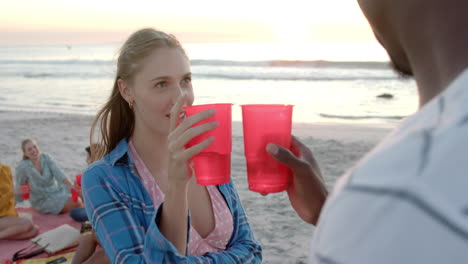 Young-Caucasian-woman-sips-from-a-red-cup-at-a-beach-party,-facing-an-African-American-man