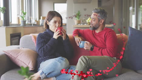 Animation-of-hearts-over-diverse-couple-sitting-on-sofa-and-drinking-coffe-at-home