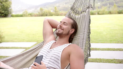 A-young-Caucasian-man-relaxes-in-a-hammock-in-the-backyard-at-home,-smartphone-in-hand