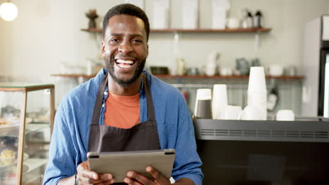 A-cheerful-African-American-male-barista-holds-a-tablet-in-a-bright-café-setting