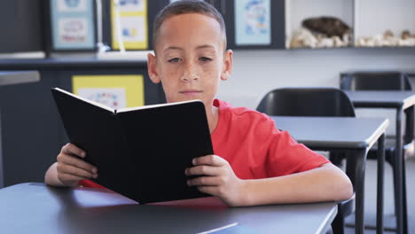 Biracial-boy-with-freckles-is-reading-a-book-in-a-classroom-at-school