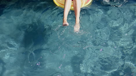 Die-Beine-Eines-Teenager-Mädchens-Baumeln-Von-Einem-Gelben-Schwimmkörper-Ins-Klare,-Blaue-Poolwasser
