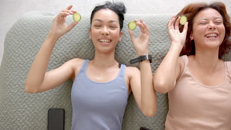 Two-female-friends-are-lying-down-with-slices-of-cucumber-on-their-eyes,-smiling-joyfully