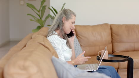 Caucasian-woman-talking-on-phone,-looking-at-laptop-in-living-room