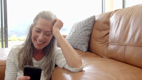 Caucasian-woman-with-grey-hair-is-sitting-on-couch,-looking-at-a-smartphone