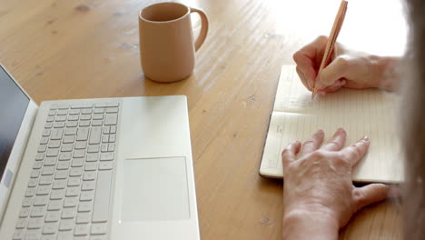 Reife-Frau-Schreibt-In-Notizbuch-Neben-Laptop-Auf-Holztisch