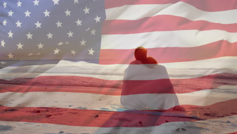 Animación-De-La-Bandera-Estadounidense-Sobre-Una-Feliz-Pareja-Diversa-Envuelta-En-Una-Alfombra-Abrazándose-Sentada-En-La-Playa-Al-Atardecer