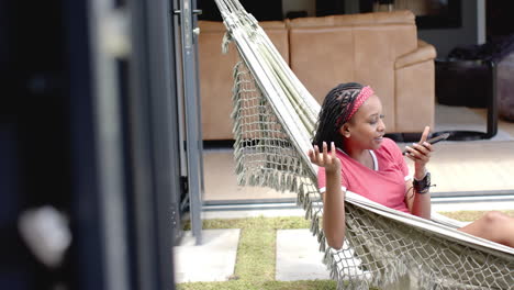 African-American-young-woman-relaxing-in-hammock,-looking-at-smartphone