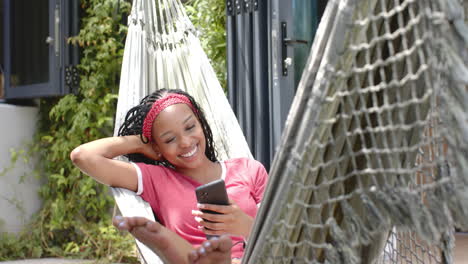 Mujer-Joven-Afroamericana-Relajándose-En-Una-Hamaca,-Sosteniendo-Un-Teléfono-Inteligente