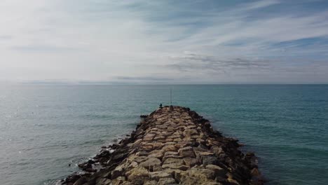 Muelle-Rocoso-Que-Se-Extiende-Hacia-El-Tranquilo-Mar-Azul-Bajo-Un-Cielo-Parcialmente-Nublado-En-Sitges,-España