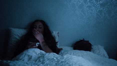 Woman-scrolls-through-social-media-and-yawns-while-sitting-in-bed-at-night