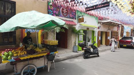 Bunter-Obstkarren-Auf-Einer-Lebhaften-Straße-In-Getsemani,-Cartagena,-Mit-Dekorativen-Flaggen-Und-Lokalen-Geschäften