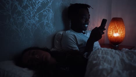 Young-man-smiles-and-looks-at-smart-phone-while-laying-in-bed-at-night