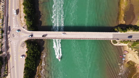 Cars-drive-over-Still-Bay-bridge-with-motorboat-passing-under,-aerial-top-down
