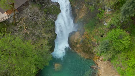 Eine-Langsame-Aufwärtsbewegung-Der-Kamera-Zeigt-Den-Sum-Wasserfall-In-Slowenien-Inmitten-Grüner-Natur,-Dessen-Wasser-In-Einen-Türkisfarbenen-Teich-Und-Eine-Holzbrücke-Neben-Einem-Haus-Fällt