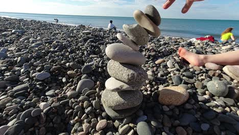 Tower-is-been-built-by-balancing-pebble-stones-on-top-of-each-other-until-they-collapse-on-background-we-can-see-various-people-and-children-feet