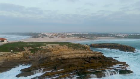 Baleal-Village-Residential-area-on-top-of-hill,-aerial-tracking-shot-towards-waves-crashing-on-the-rocky-coast