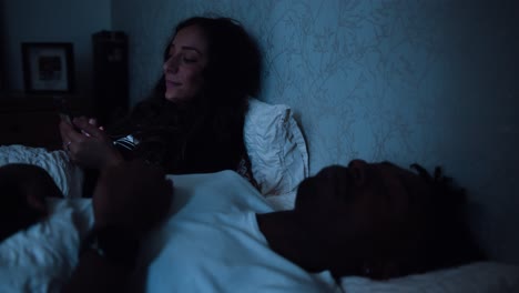 Woman-scrolls-on-her-phone-while-her-partner-sleeps-in-bed-next-to-her-late-at-night
