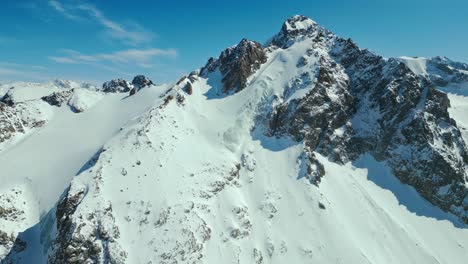 Hermoso-Cielo-Azul,-Erosión-Rocosa,-Cumbre-De-La-Montaña,-Nieve-A-La-Deriva,-Antena