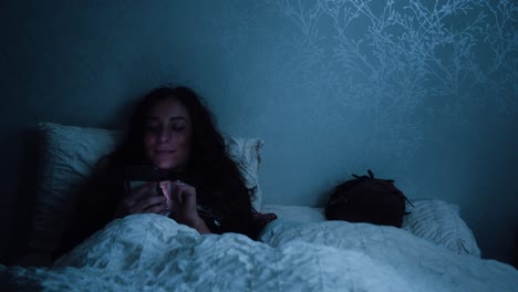 Woman-smiles-at-her-phone-whilst-scrolling-through-social-media-late-at-night