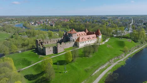 Aerial-establishing-view-of-Bauska-Medieval-Castle-and-ruins,-Musa-and-Memele-rivers-next-to-the-castle,-sunny-spring-day,-wide-drone-shot-moving-forward,-tilt-down