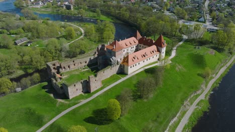 Aerial-establishing-view-of-Bauska-Medieval-Castle-and-ruins,-Musa-and-Memele-rivers-next-to-the-castle,-sunny-spring-day,-wide-drone-shot-moving-backward,-camera-tilt-up