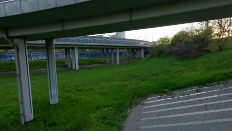 Drohnenaufnahme-Eines-Autos-Auf-Einer-Hochbrücke-Mit-Grünem-Gras-Darunter-Und-Einer-Stadt-Im-Hintergrund