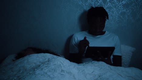 Man-uses-tablet-device-sitting-in-bed-feeling-tired-but-staying-up-late-at-night