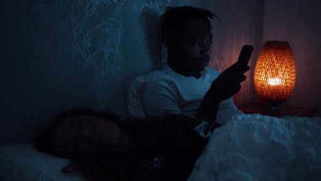 Man-uses-smart-phone-at-late-at-night-while-partner-sleeps-in-the-bed-next-to-him