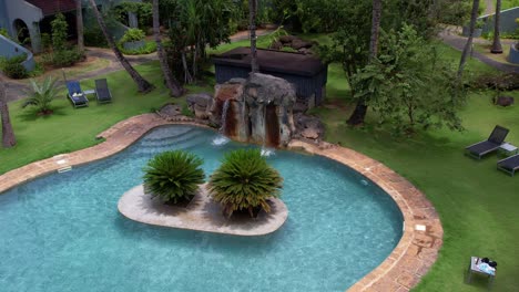 Resort-swimming-pool-with-waterfall-surrounded-by-Lush-vegetation,-Aerial-Orbit-shot