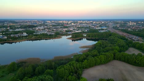 Panorama-Al-Amanecer,-Vuelo-De-Drones-Sobre-El-Bosque-Y-El-Lago,-Con-Edificios-Del-Distrito-Industrial-Visibles-Al-Fondo