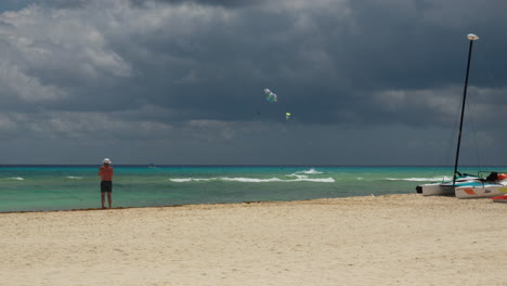 Ein-Mann-Steht-An-Einem-Strand-In-Der-Nähe-Des-Ozeans-Und-Blickt-Auf-Gleitschirmflieger-Vor-Einem-Dunklen,-Dramatischen-Wolkenhimmel