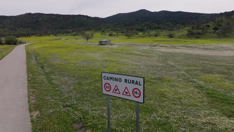 flight-with-a-drone-visualizing-a-meadow-full-of-white-and-yellow-flowers-with-a-sign-indicating-a-Rural-Road-and-its-regulations,-said-road-is-on-one-side-we-see-a-background-of-mountains-Avila