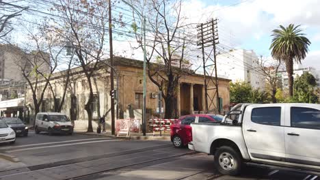Los-Autos-Modernos-Cruzan-Una-Antigua-Estación-De-Tren-Con-Una-Casa-Colonial-En-La-Ciudad-De-Buenos-Aires,-Argentina,-Un-Día-Panorámico-Otoñal-Con-Vegetación-Local-En-El-Barrio-De-Flores.