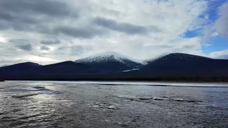 Montañas-Cubiertas-De-Nieve-Bajo-Un-Cielo-Nublado,-Con-Un-Río-Parcialmente-Congelado-En-Primer-Plano