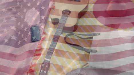 Animation-of-american-flag-and-light-spots-over-guitar,-smartphone-and-bag-on-beach-towels-in-sun