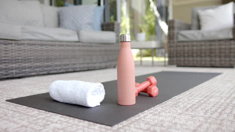 A-yoga-mat,-white-towel,-pink-water-bottle,-and-dumbbells-rest-on-patio